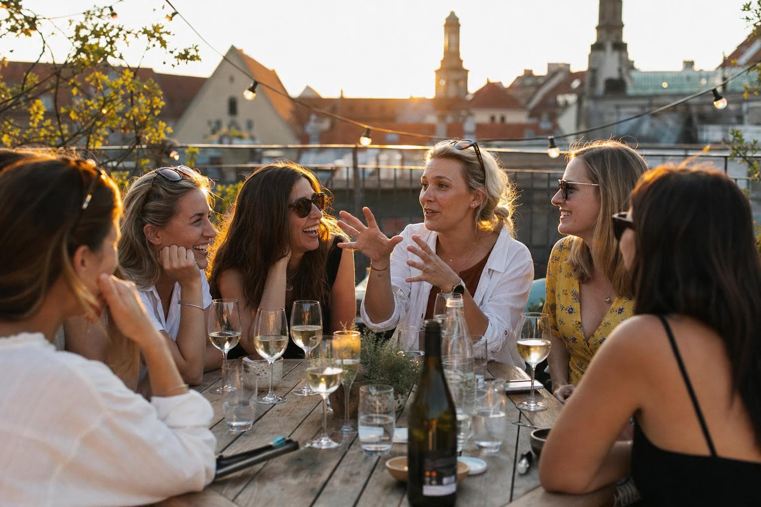 A hen party laughing over prosecco at a rooftop table at golden hour